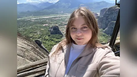 Smiling young woman in a beige jacket enjoying a scenic mountain view at a high altitude lookout.