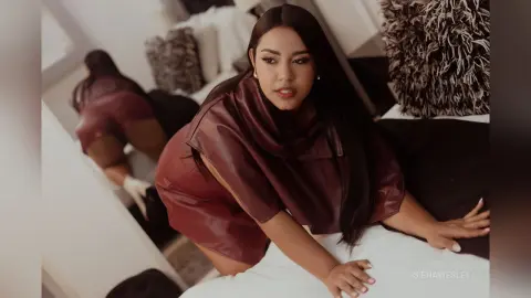 SienaWesley wears a dark maroon leather outfit while leaning forward on a white bedsheet.