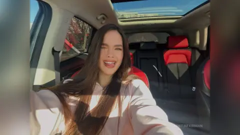Smiling young woman in a pink sweatshirt sitting in a car with a sunroof on a sunny day.