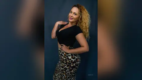 Smiling blonde woman in a black v-neck top and leopard print skirt posing against a dark studio background.