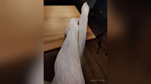 RubyAttwood rests her legs on a wooden table while wearing white lace stockings.