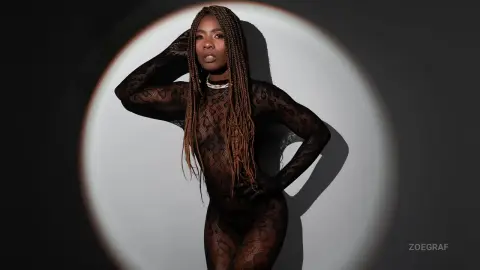 Elegant Black woman with long braids in a sheer black lace bodysuit posing against a dramatic spotlight background.