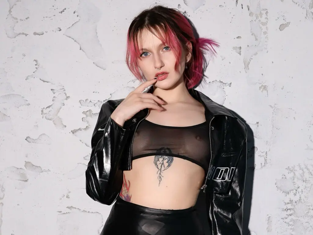 Edgy woman with pink hair in a black leather jacket and sheer top posing against a textured white wall.