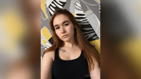 JaneBarbrick poses in a black tank top with her long brown hair down against a yellow and grey patterned background.