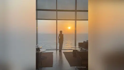 Silhouette of a woman standing by a large window overlooking a golden ocean sunset in a modern luxury room.
