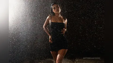 Moody portrait of a woman in a black dress standing under falling rain against a dark background.