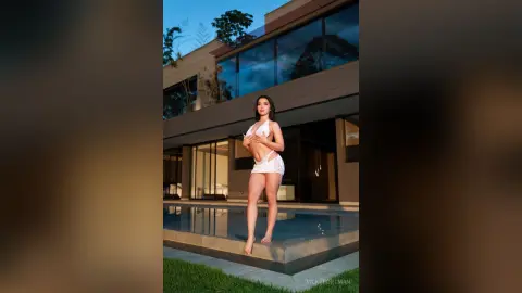 VickyPortman stands poolside in a white lace teddy, showing her bare legs and cleavage against a modern glass house.