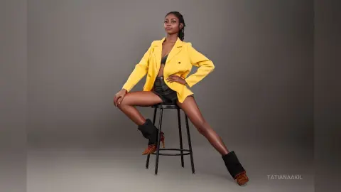 Stylish woman in a bright yellow blazer and black shorts sitting on a stool against a grey studio background.