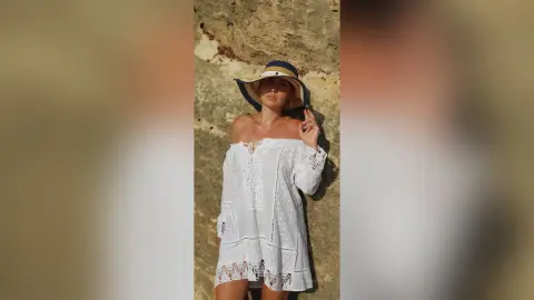 AryaSin poses in a white lace off-the-shoulder dress and a wide-brimmed sun hat against a stone wall.