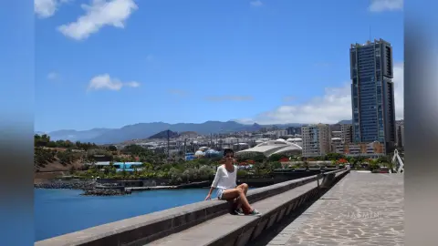 Young woman in a white t-shirt and denim shorts relaxing on a stone ledge by a blue lagoon under a sunny sky.
