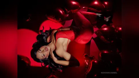 NohomiCambel poses in a red bodysuit with her long black hair flowing against a dark red background.