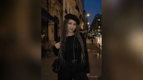 Woman with long braids in a black outfit and beret walking on a city street at night.
