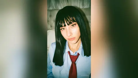 Young woman with black bangs wearing a light blue button-down shirt and red tie in a moody indoor setting.