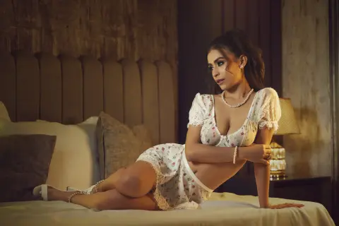 Elegant woman in white lace lingerie sitting on a bed in a moody, dimly lit bedroom setting.