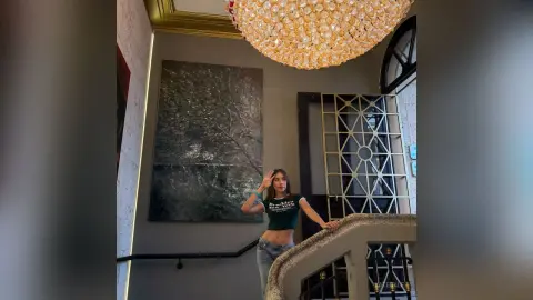 KateGale poses in a black t-shirt and light blue jeans, standing near a staircase and a large painting under a golden chandelier.