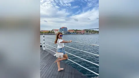 Stylish woman in white top and denim shorts posing on a wooden pier overlooking a coastal town under a blue sky.