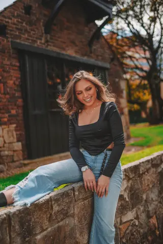 Smiling young woman in a black long-sleeve top and light blue jeans sitting on a stone wall outdoors.