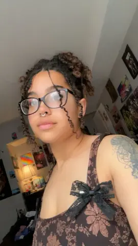 PrincessBiancaHoney wearing black glasses and a brown floral sundress with a black lace bow, showing her curly hair and shoulder tattoo.