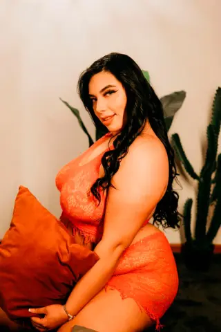 Curvy woman in orange lace loungewear sitting by indoor plants in a warm, cozy setting.
