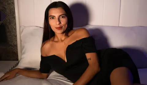 Smiling woman in a black off-the-shoulder dress relaxing on a white bed in soft indoor lighting.