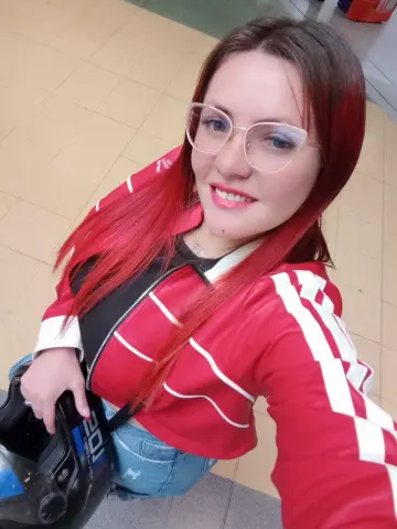Smiling woman with red hair wearing a red and white track jacket in a casual indoor setting.