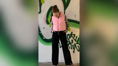 Young woman in a pink puffer vest and black wide-leg trousers standing against a graffiti wall.