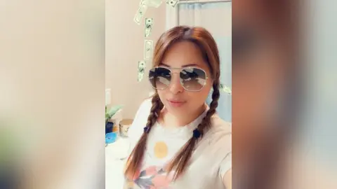 DaniReed wears sunglasses and has her hair in two long pigtail braids while wearing a white t-shirt.