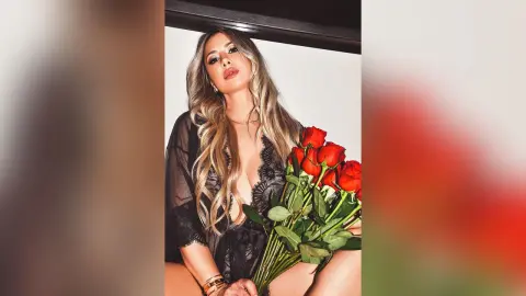 Glamorous blonde woman in a black lace robe holding a bouquet of red roses in a romantic indoor setting.