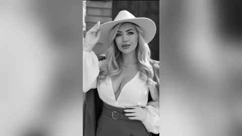 Anelise poses wearing a white hat and a low-cut white blouse that shows her cleavage, looking directly at the camera.