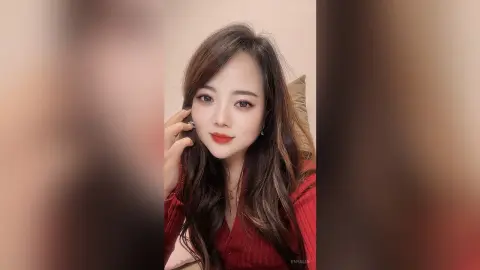 EnyaLin poses with a hand to her face, wearing a red top with her long brown hair styled in waves.