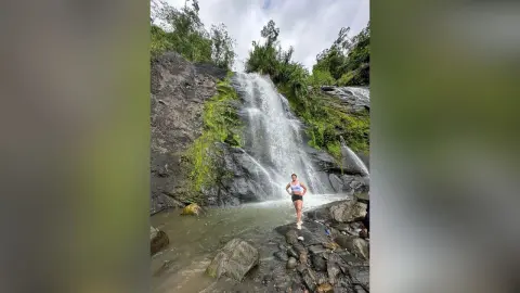 Woman in white tank top and black shorts standing by a scenic waterfall in a lush tropical forest.