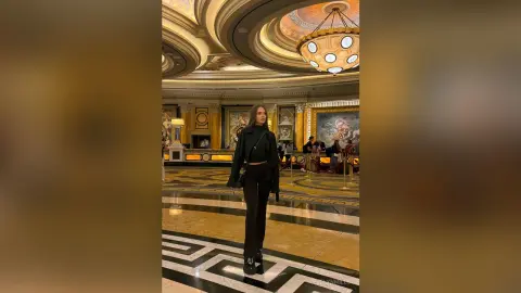 EvelynMills stands in a grand, gold-accented hall wearing a black long-sleeved crop top and matching black trousers.