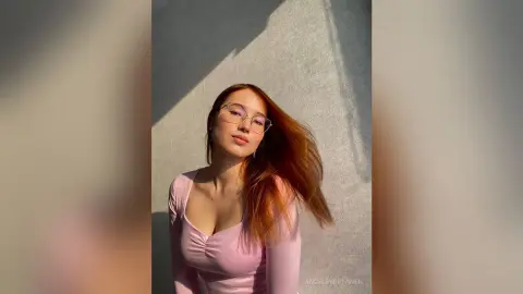 Redheaded woman in a pink long-sleeve top wearing glasses, posing in warm sunlight against a grey wall.