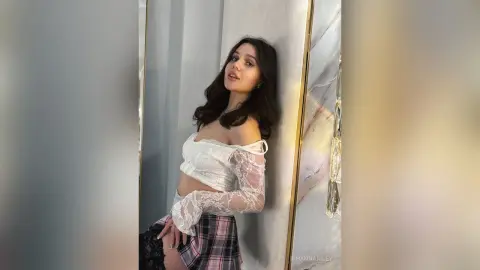 Young woman in a white lace off-the-shoulder top and plaid skirt posing elegantly against a marble wall.