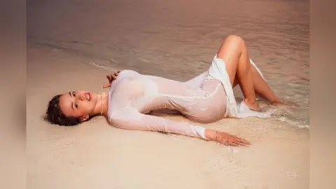 Elie lies on a sandy beach wearing a wet, sheer white long-sleeve top that shows her nipples through the fabric.
