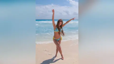 AndreaKalo stands on a sandy beach wearing a yellow and blue patterned bikini, posing with her arms raised against the ocean waves and blue sky.