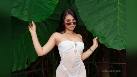 Stylish woman in a white strapless polka dot dress and sunglasses posing against large green tropical leaves.