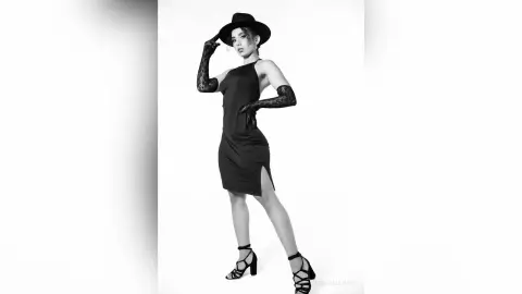 Elegant woman in a black cocktail dress and wide-brimmed hat posing in a sophisticated black and white studio portrait.
