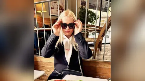 MilenaBia wears dark sunglasses and a navy blue blazer over a white top while sitting at a wooden table outdoors.