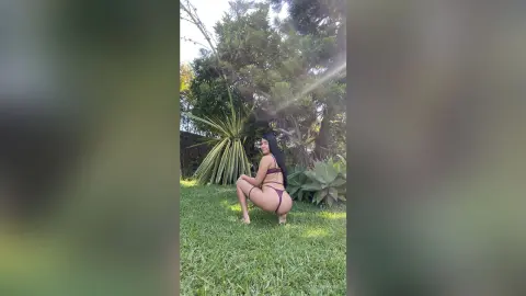 Young woman in a dark bikini posing on a sunny green lawn surrounded by tropical plants and trees.
