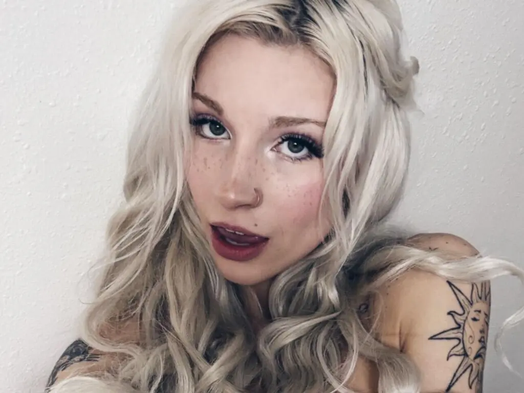 Close-up portrait of a blonde woman with freckles and tattoos posing against a white wall with a soft mood.