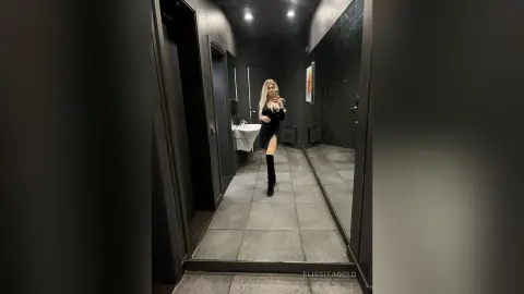 Blonde woman in a chic black mini dress and tall boots posing in a modern dark hallway mirror.