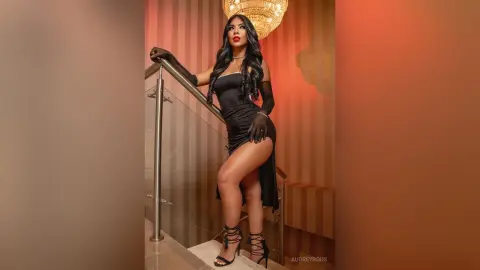 Elegant woman in a black cocktail dress and gloves posing on a modern staircase with warm lighting.