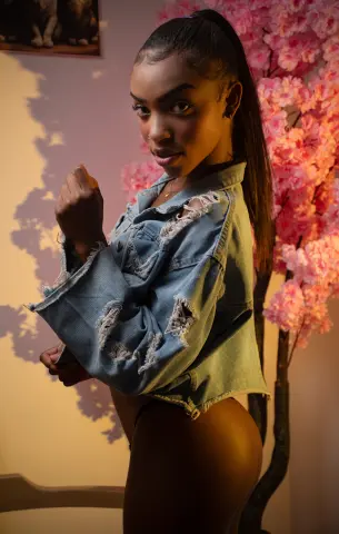 CahitlynBel poses in a ripped denim jacket, looking over her shoulder with her hair in a high ponytail next to pink cherry blossoms.