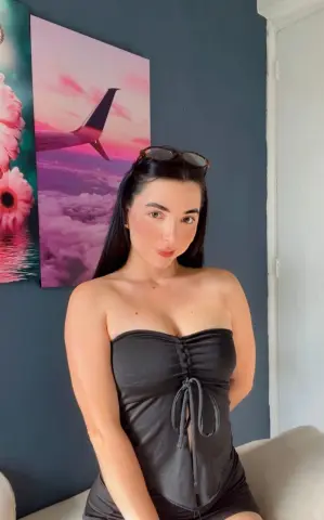 Young woman in a black strapless romper posing indoors against a pink sunset landscape painting.