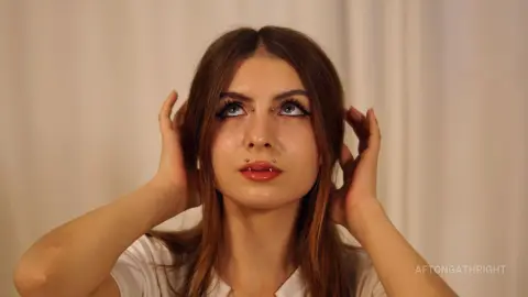 Young woman with facial piercings and long brown hair posing in a white shirt against a neutral background.