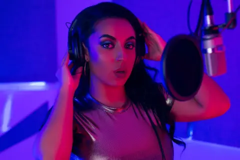 Woman in a metallic top singing into a studio microphone under moody blue and pink neon lighting.