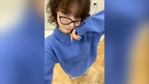 Young woman with glasses wearing a cozy blue knit sweater posing in a bright room.