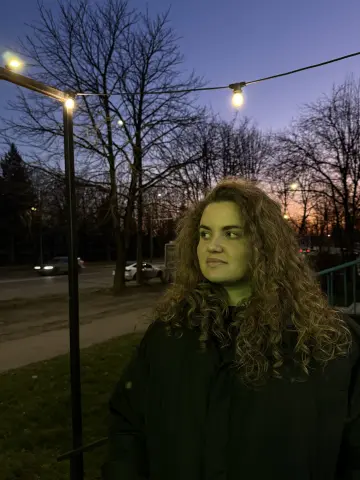 Curly-haired woman in a black jacket standing outdoors under string lights at twilight.