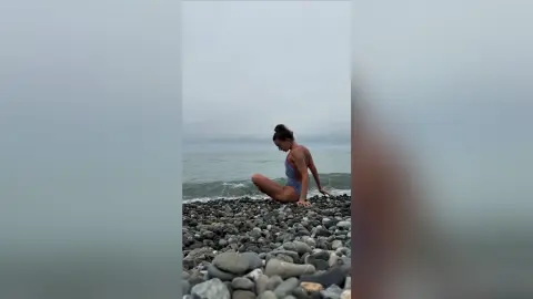 Young woman in a red swimsuit sitting on a rocky beach under a moody, overcast sky.